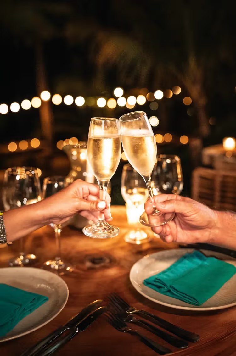 Two glasses of sparkling wine raised in a toast at a candlelit table under string lights